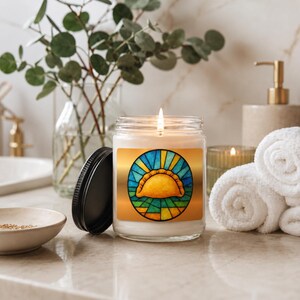 May include: A clear glass jar candle with a lit wick and a black lid. The label features a circular stained glass design in shades of blue, green, and yellow, depicting a golden-brown empanada centered within the geometric pattern.