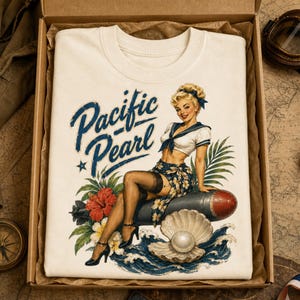 May include: A cream-colored t-shirt featuring a graphic print of a blonde pin-up figure in a sailor outfit sitting on a torpedo, surrounded by tropical flowers, waves, and a pearl in a shell, with the blue text Pacific Pearl printed above.