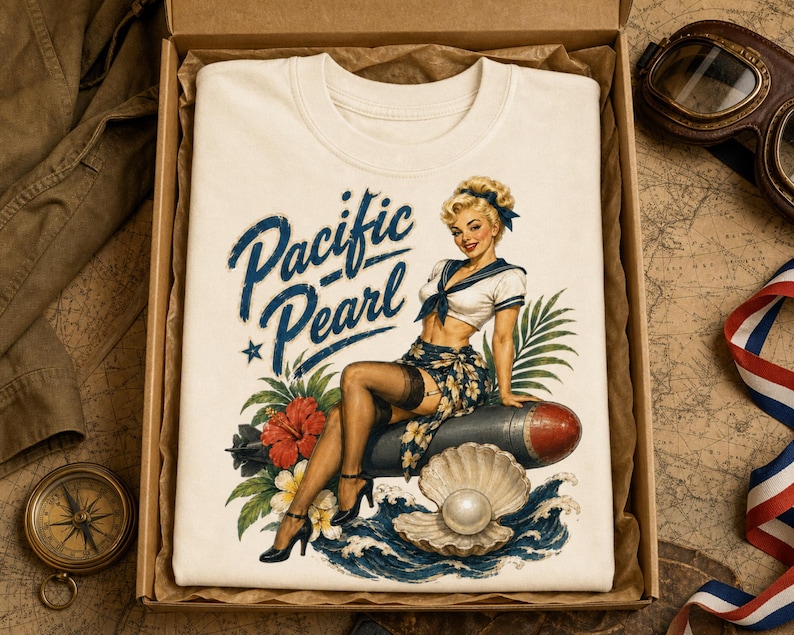 May include: A cream-colored t-shirt featuring a graphic print of a blonde pin-up figure in a sailor outfit sitting on a torpedo, surrounded by tropical flowers, waves, and a pearl in a shell, with the blue text Pacific Pearl printed above.
