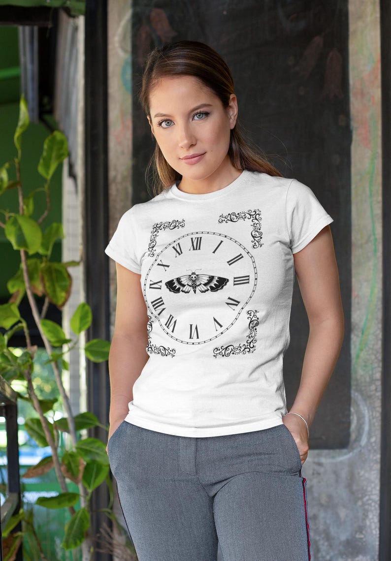 May include: White t-shirt featuring a black clock design with Roman numerals and a moth graphic at the center. The clock is framed by decorative black flourishes. The model is wearing gray pants and standing in front of a textured background.