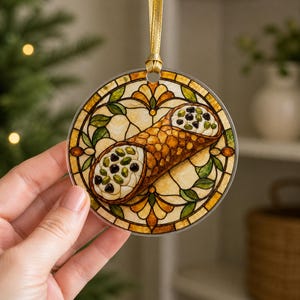 May include: A circular hanging ornament featuring a stained glass style illustration of a cannoli pastry. The design includes a golden-brown shell filled with white cream, dark chocolate chips, and green pistachios, framed by amber and green glass patterns.