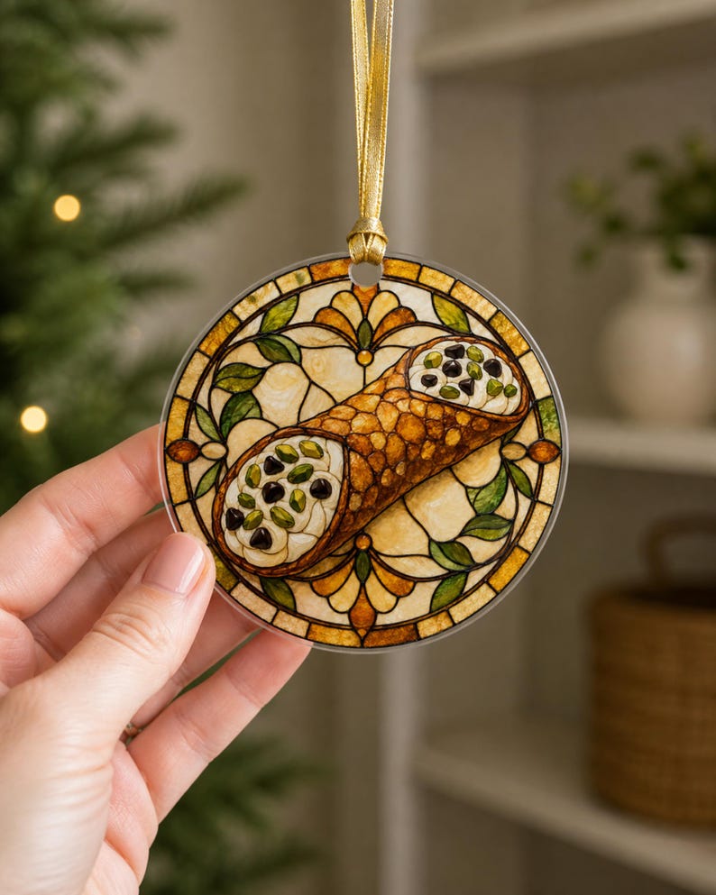 May include: A circular hanging ornament featuring a stained glass style illustration of a cannoli pastry. The design includes a golden-brown shell filled with white cream, dark chocolate chips, and green pistachios, framed by amber and green glass patterns.