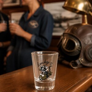 May include: A clear glass shot glass featuring a black and white graphic of a cartoon cat wearing goggles and riding a bomb, with the text Lucky 13 printed in black script above the illustration. The glass has a thick base and straight sides.