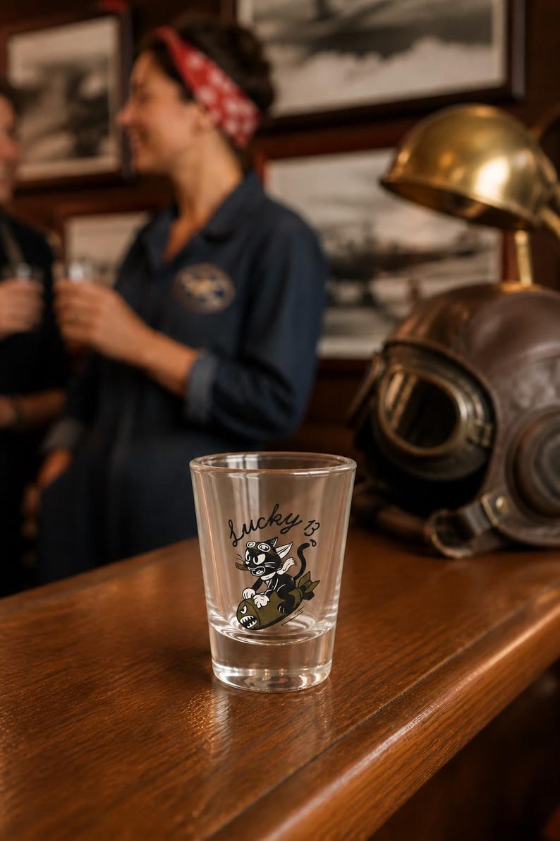 May include: A clear glass shot glass featuring a black and white graphic of a cartoon cat wearing goggles and riding a bomb, with the text Lucky 13 printed in black script above the illustration. The glass has a thick base and straight sides.