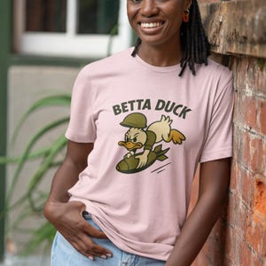 May include: A pale pink t-shirt featuring the words "BETTA DUCK" and a cartoon duck riding a bomb. The duck wears a military helmet and uniform. The person in the image is wearing blue jeans.