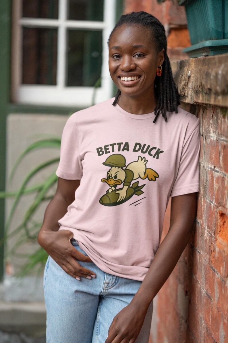 May include: A pale pink t-shirt featuring the words "BETTA DUCK" and a cartoon duck riding a bomb. The duck wears a military helmet and uniform. The person in the image is wearing blue jeans.
