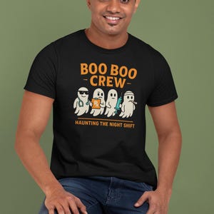 May include: Black t-shirt with the text "BOO BOO CREW" in orange. The shirt features a graphic of four cartoon ghosts, each with a medical-themed accessory. The phrase "HAUNTING THE NIGHT SHIFT" is printed below the ghosts.