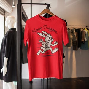 May include: A red t-shirt with the words "Hare Trigger" and a cartoon rabbit running with a football. The shirt is hanging on a wooden hanger in a clothing store.