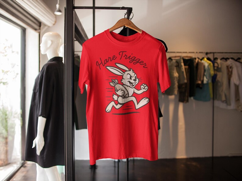 May include: A red t-shirt with the words "Hare Trigger" and a cartoon rabbit running with a football. The shirt is hanging on a wooden hanger in a clothing store.