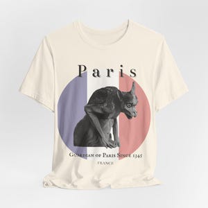 May include: Cream-colored t-shirt featuring a black and white gargoyle illustration. The design includes a French flag-inspired circle with the word "Paris" above the gargoyle and the text "Guardian of Paris Since 1345 France" below.