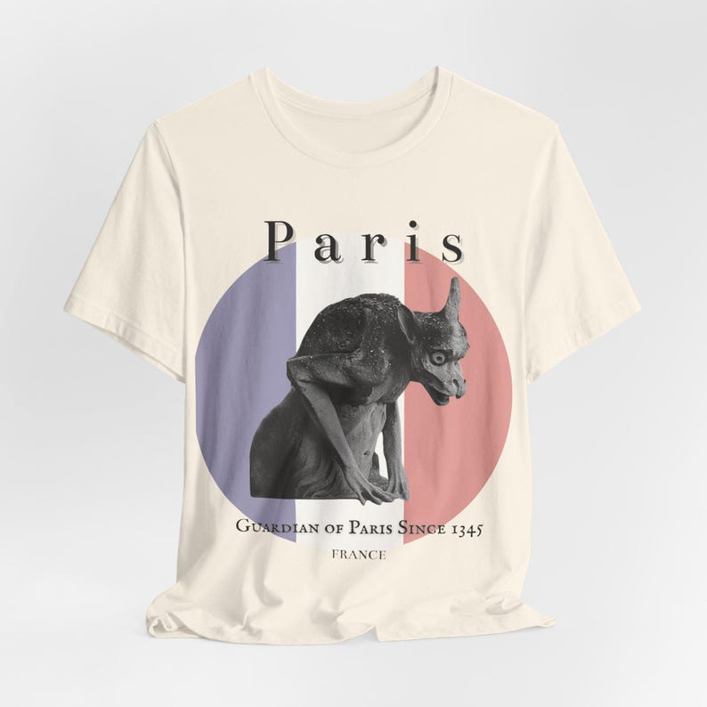 May include: Cream-colored t-shirt featuring a black and white gargoyle illustration. The design includes a French flag-inspired circle with the word "Paris" above the gargoyle and the text "Guardian of Paris Since 1345 France" below.
