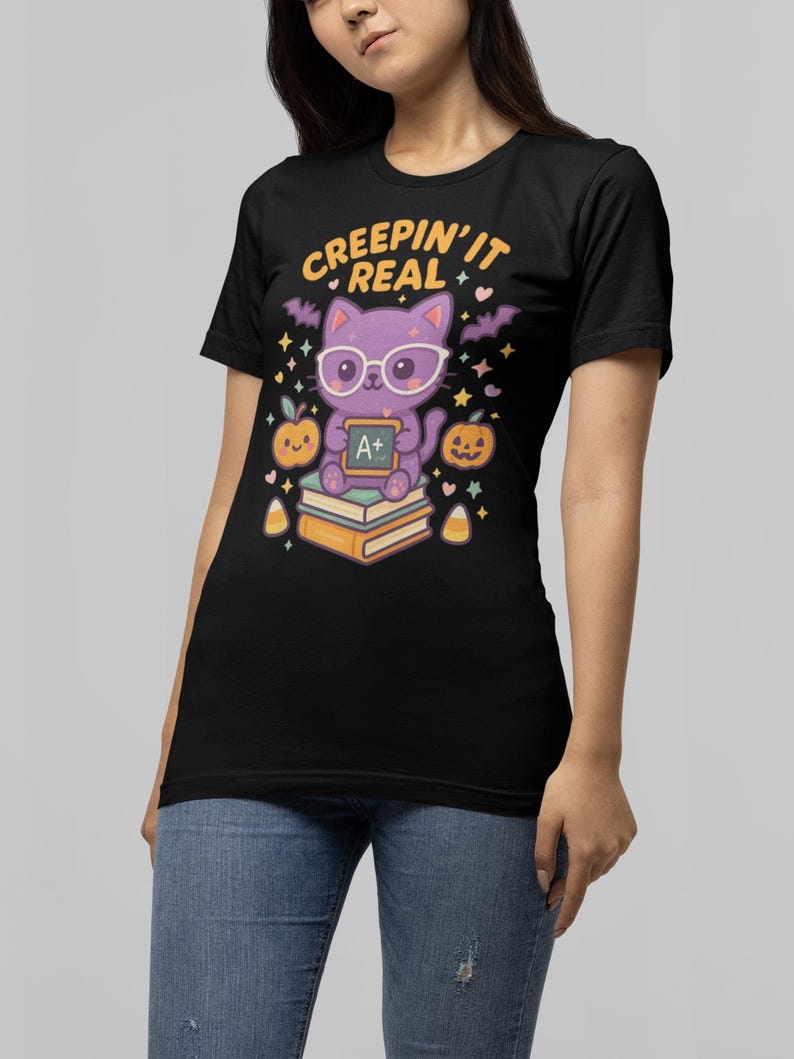 May include: Black t-shirt with a Halloween-themed graphic. The design features a purple cat wearing glasses and holding an A+ sign, with the text "CREEPIN' IT REAL". The cat is sitting on books, with pumpkins and bats.