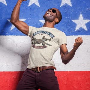 May include: A light beige short-sleeve t-shirt featuring a graphic of a vintage military aircraft. The text reads America 1776-2026 and 250 Years. The shirt is paired with dark maroon trousers and a brown leather belt with a metal buckle.