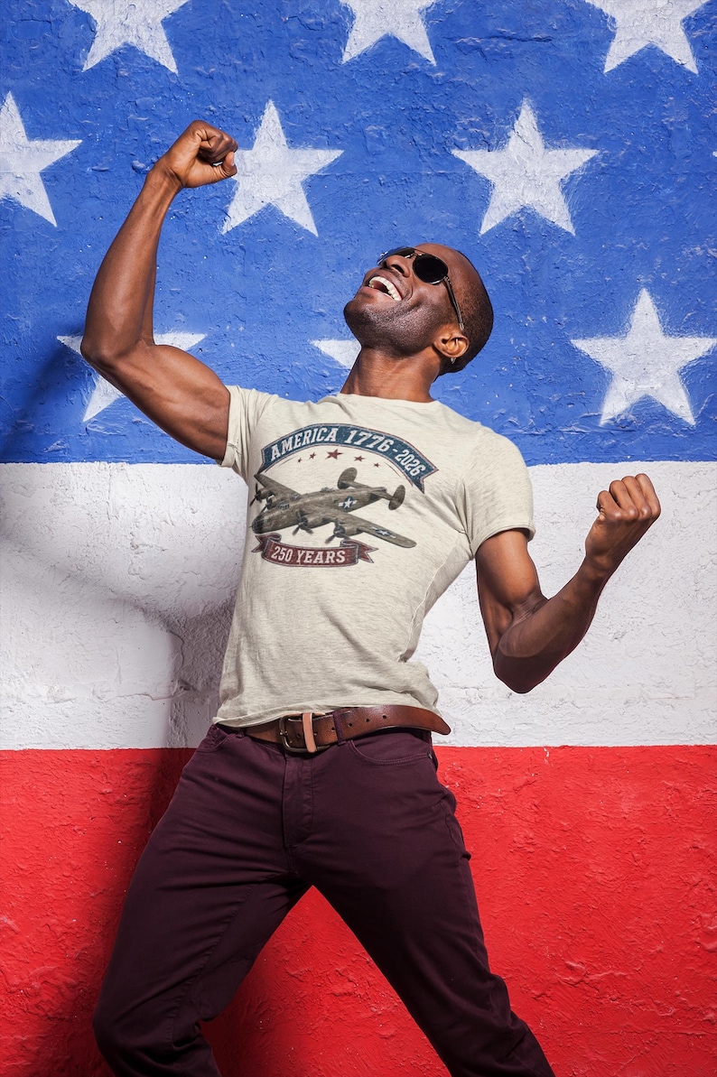 May include: A light beige short-sleeve t-shirt featuring a graphic of a vintage military aircraft. The text reads America 1776-2026 and 250 Years. The shirt is paired with dark maroon trousers and a brown leather belt with a metal buckle.