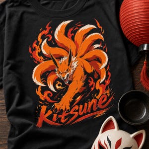 K&ouml;nnte beinhalten: Ein schwarzes T-Shirt mit einer grafischen Illustration eines orangefarbenen Fuchses mit mehreren Schw&auml;nzen, umgeben von stilisierten orangefarbenen und roten Flammen. Das Wort Kitsune ist in einer fetten Pinselstrich-Schrift in Rot und Orange gedruckt.