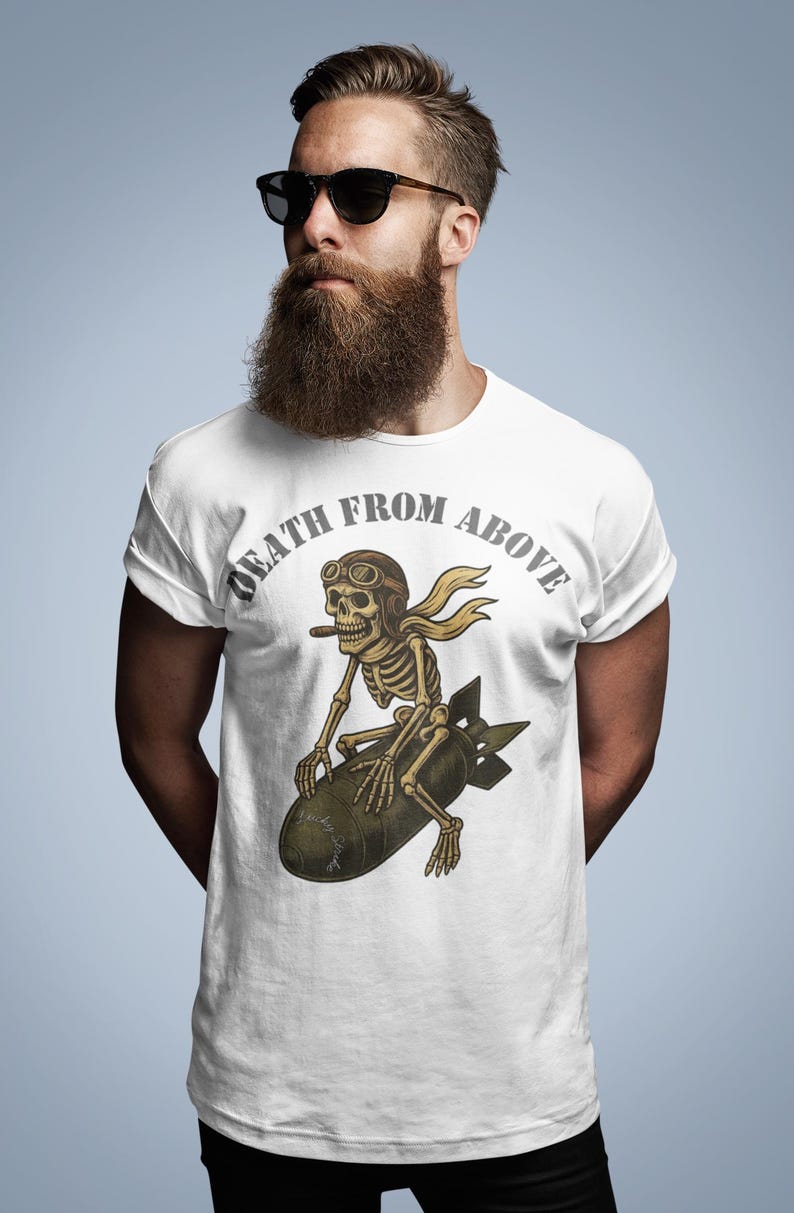 May include: White t-shirt featuring a skeleton wearing aviator goggles and a scarf, riding a bomb. The text "DEATH FROM ABOVE" is arched above the skeleton. The design is in shades of brown and tan.