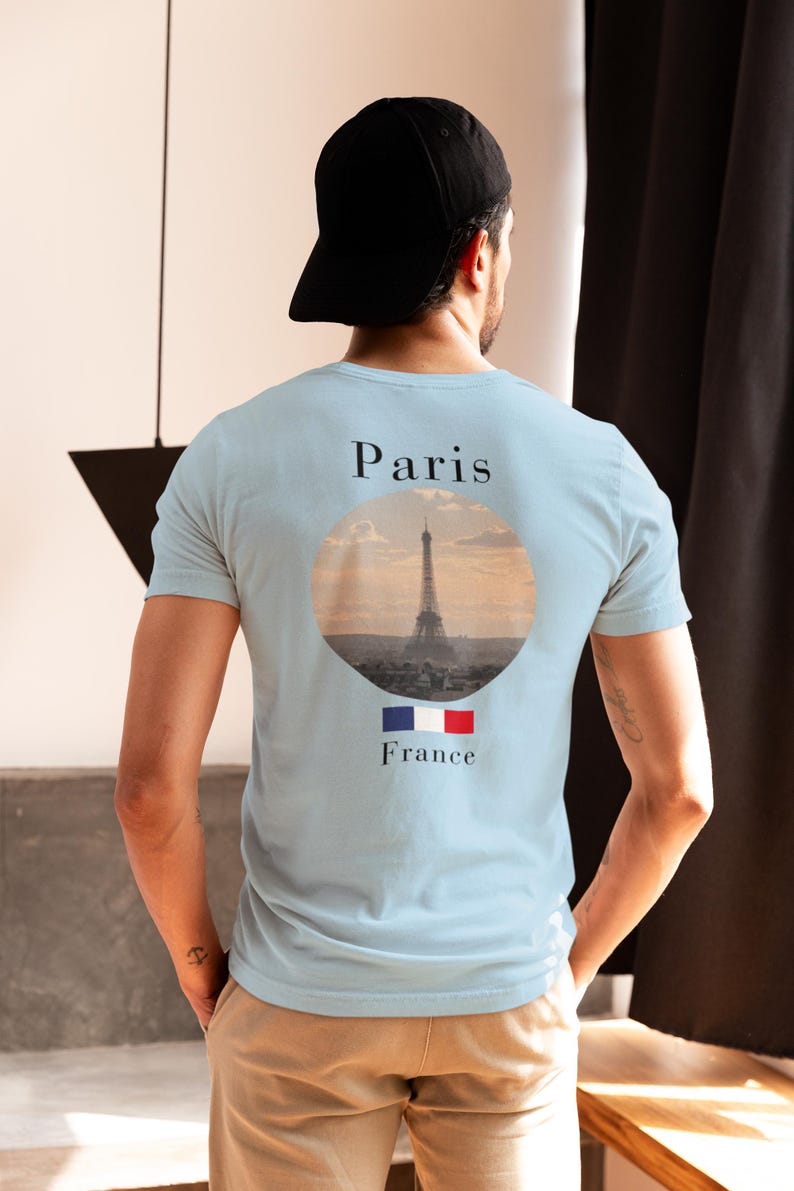 May include: Light blue t-shirt with a graphic on the back featuring a circular image of the Eiffel Tower in Paris, France. The text 'Paris' is above the image, and 'France' is below, with the French flag. The shirt is worn by a person with a black cap.