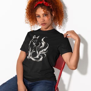 May include: A black short-sleeved t-shirt featuring a white ink-style illustration of a multi-tailed fox sitting in profile. The graphic includes a small red square stamp detail on the left side of the fox, set against a solid black fabric background.