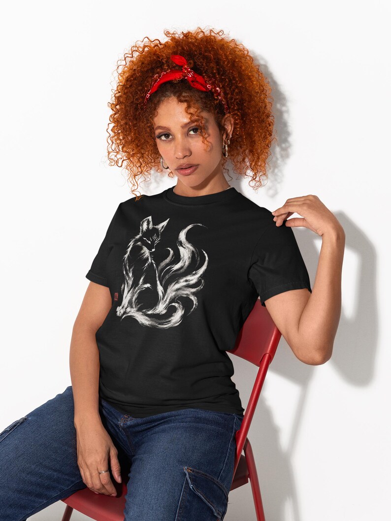 May include: A black short-sleeved t-shirt featuring a white ink-style illustration of a multi-tailed fox sitting in profile. The graphic includes a small red square stamp detail on the left side of the fox, set against a solid black fabric background.