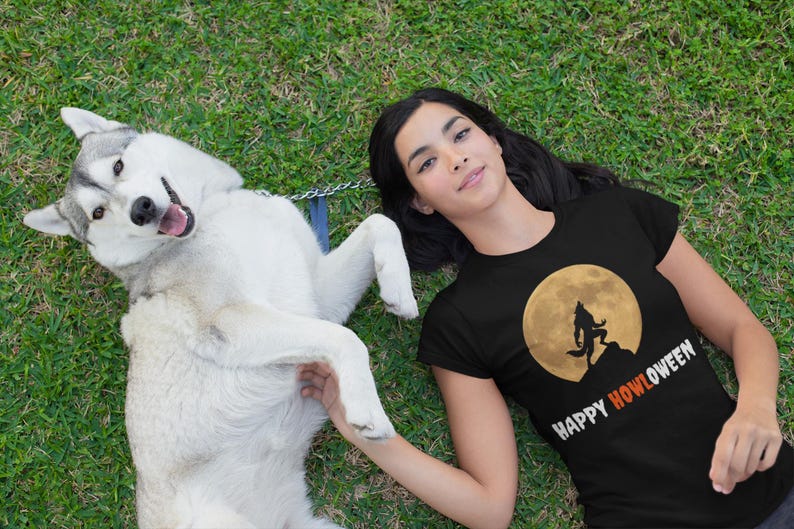 May include: A black t-shirt with a Halloween design featuring a werewolf silhouette against a full moon, with the text "HAPPY HOWLOWEEN". A white and gray husky dog is lying on the grass.