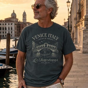 May include: A dark gray short-sleeve t-shirt featuring a white graphic print of the Rialto Bridge in Venice, Italy. The text reads VENICE ITALY, LAGUNA DI VENEZIA, La Serenissima, and EST. 421 AD, depicting a gondola and birds in the scene.