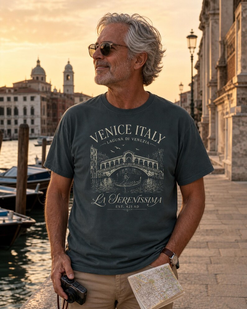 May include: A dark gray short-sleeve t-shirt featuring a white graphic print of the Rialto Bridge in Venice, Italy. The text reads VENICE ITALY, LAGUNA DI VENEZIA, La Serenissima, and EST. 421 AD, depicting a gondola and birds in the scene.