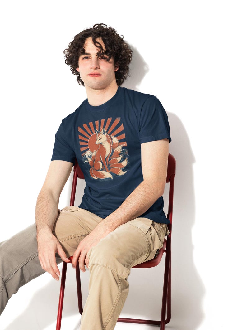 May include: A navy blue short-sleeve t-shirt featuring a graphic print of a multi-tailed fox sitting in front of a large red sun with radiating beams. The design includes stylized waves at the base of the fox, rendered in shades of orange, white, and dark blue.