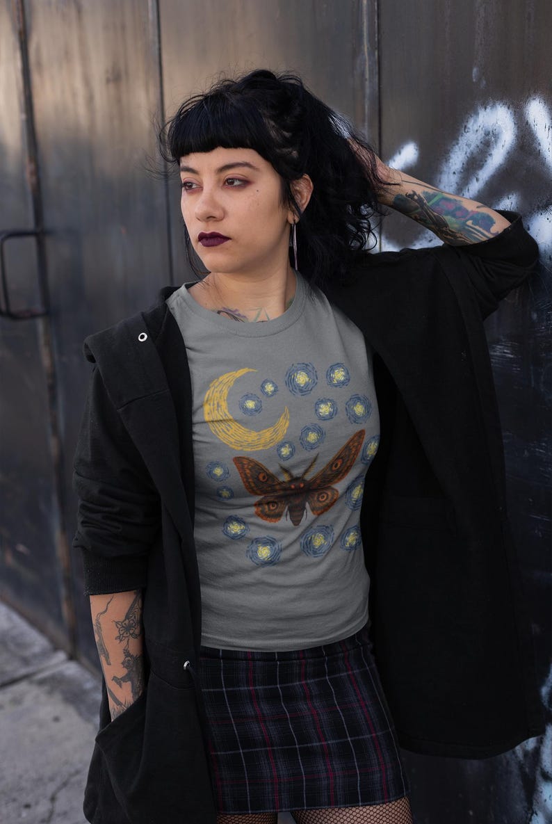 May include: Gray t-shirt featuring a moth design with a crescent moon and starry night sky. The moth is brown with orange accents. The shirt is paired with a plaid skirt and black jacket.