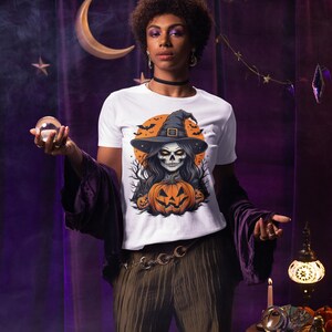 May include: White t-shirt with a Halloween graphic. The design shows a skull in a witch hat, a carved pumpkin, and bats against an orange backdrop. The shirt is a short-sleeved crew neck.