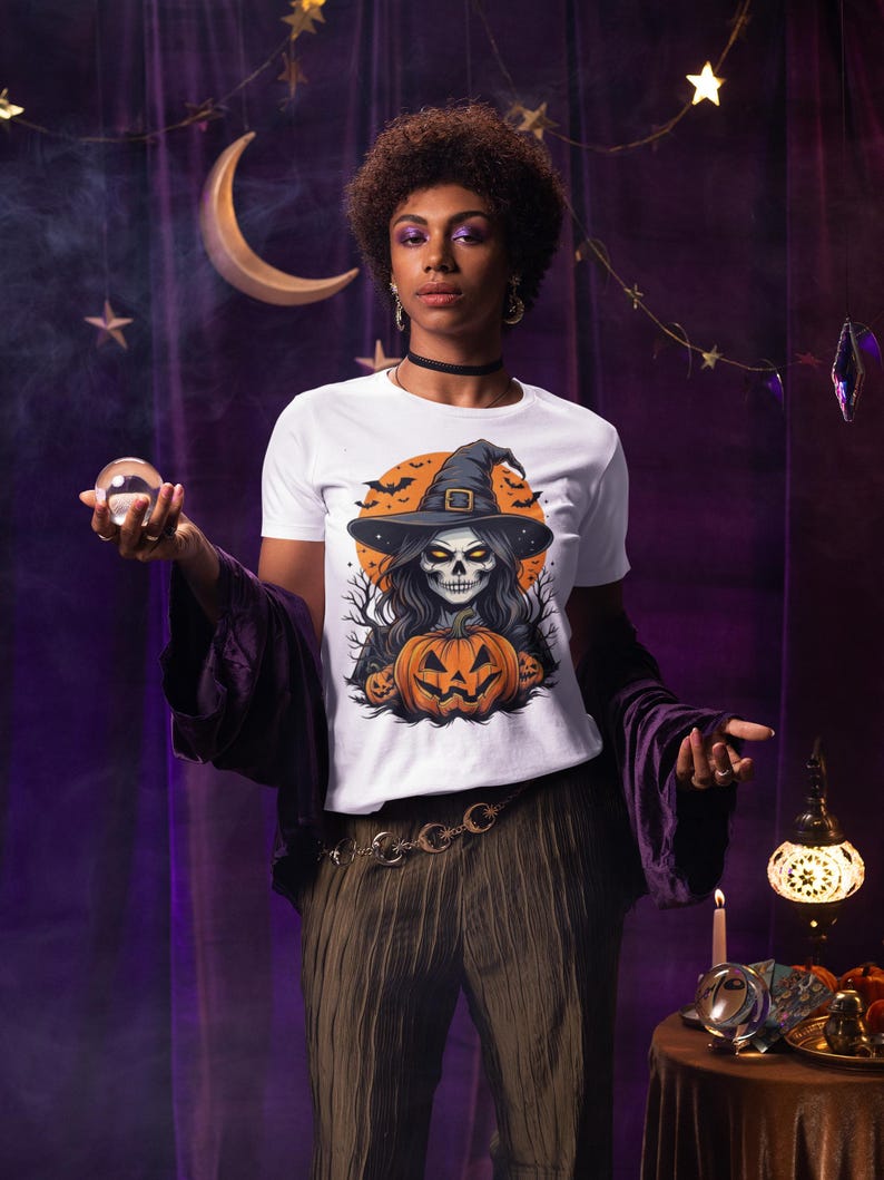 May include: White t-shirt with a Halloween graphic. The design shows a skull in a witch hat, a carved pumpkin, and bats against an orange backdrop. The shirt is a short-sleeved crew neck.