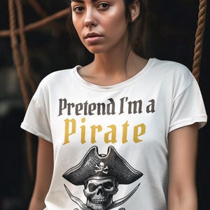 May include: White t-shirt with the text "Pretend I'm a Pirate" in black and gold lettering. Below the text is a black and white illustration of a pirate skull wearing a hat with crossed swords. The shirt is short-sleeved.