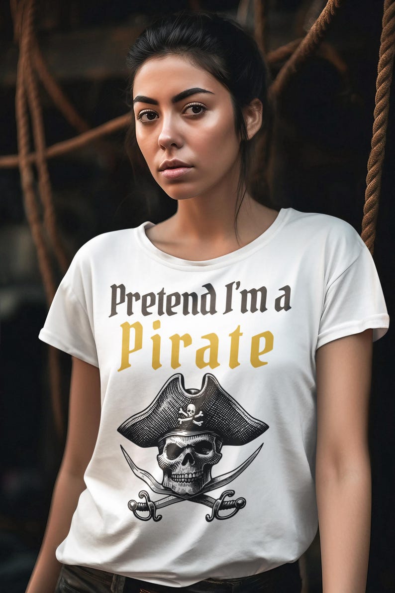 May include: White t-shirt with the text "Pretend I'm a Pirate" in black and gold lettering. Below the text is a black and white illustration of a pirate skull wearing a hat with crossed swords. The shirt is short-sleeved.