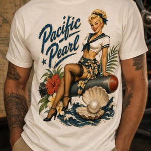 May include: A cream-colored short-sleeve t-shirt featuring a graphic print of a blonde pin-up figure in a sailor outfit sitting on a bomb, surrounded by hibiscus flowers, ocean waves, and a pearl in a shell, with the text Pacific Pearl.