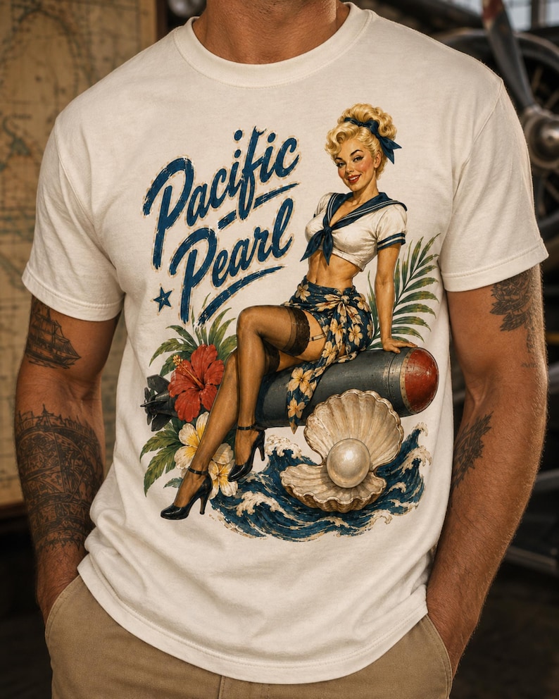 May include: A cream-colored short-sleeve t-shirt featuring a graphic print of a blonde pin-up figure in a sailor outfit sitting on a bomb, surrounded by hibiscus flowers, ocean waves, and a pearl in a shell, with the text Pacific Pearl.