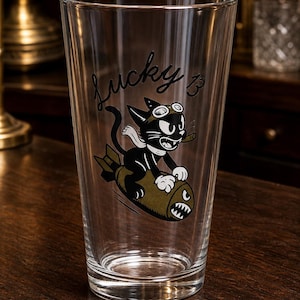 May include: A clear glass pint tumbler featuring a black and white cartoon cat wearing goggles and a scarf, riding an olive green bomb with a shark mouth graphic. The text Lucky 13 is printed in black script above the cat illustration on the glass.