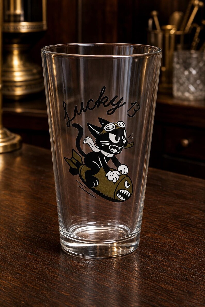 May include: A clear glass pint tumbler featuring a black and white cartoon cat wearing goggles and a scarf, riding an olive green bomb with a shark mouth graphic. The text Lucky 13 is printed in black script above the cat illustration on the glass.