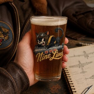 Op de afbeelding: Een helder bierglas gevuld met amberkleurig bier met een afbeelding van een zwarte kat met een vliegeniersbril die op een bom rijdt. Onder de kat staat de tekst Nine Lives in een geel lettertype met drie kleine ronde rood-witte emblemen.