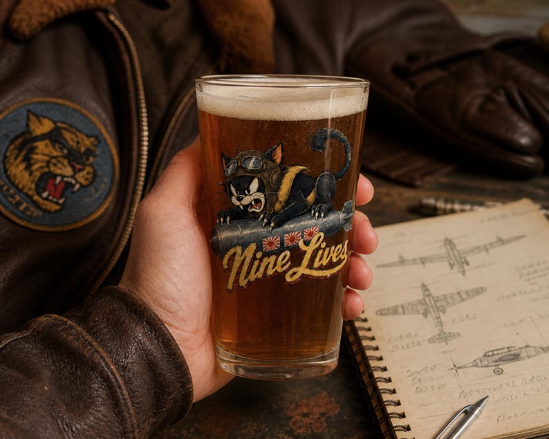 Op de afbeelding: Een helder bierglas gevuld met amberkleurig bier met een afbeelding van een zwarte kat met een vliegeniersbril die op een bom rijdt. Onder de kat staat de tekst Nine Lives in een geel lettertype met drie kleine ronde rood-witte emblemen.