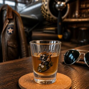 May include: A clear glass shot glass filled with an amber liquid. The glass features a graphic of a black cat wearing goggles and riding a bomb, with the text Lucky 13 written in black script above the illustration. The glass sits on a round cork coaster.
