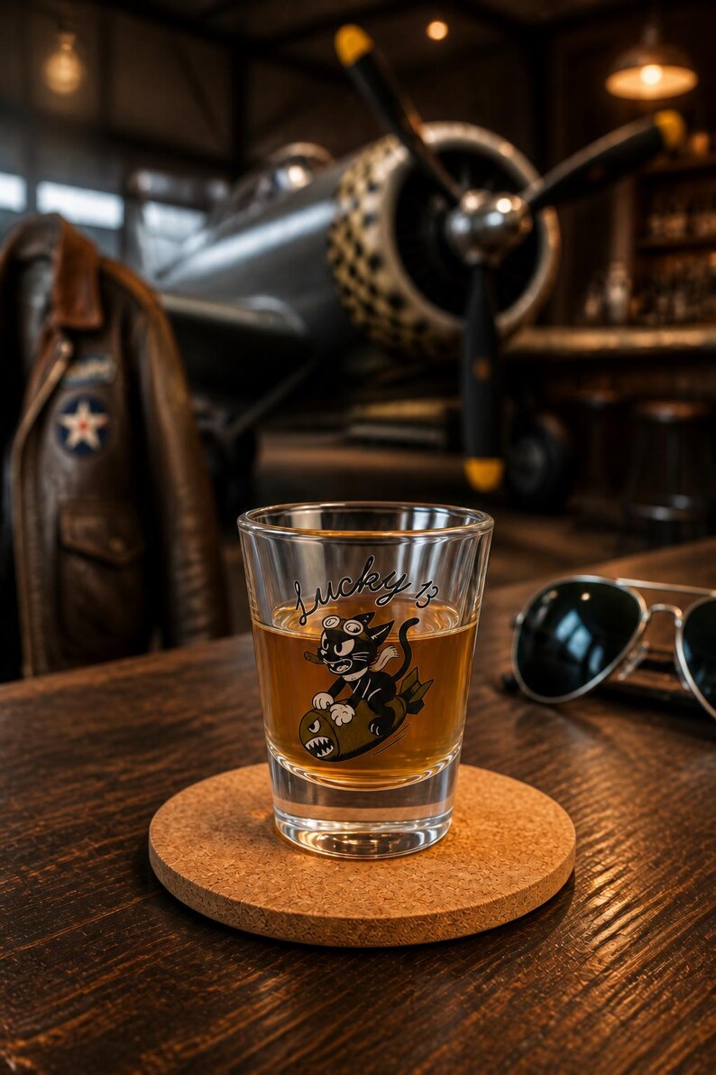 May include: A clear glass shot glass filled with an amber liquid. The glass features a graphic of a black cat wearing goggles and riding a bomb, with the text Lucky 13 written in black script above the illustration. The glass sits on a round cork coaster.