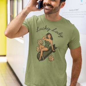 May include: An olive green short-sleeve t-shirt featuring a graphic print of a woman in a green swimsuit sitting on a dark gray bomb, surrounded by three white dice. The text Lucky Lady is printed in a dark cursive font above the illustration.