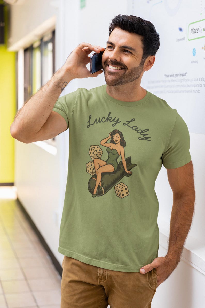 May include: An olive green short-sleeve t-shirt featuring a graphic print of a woman in a green swimsuit sitting on a dark gray bomb, surrounded by three white dice. The text Lucky Lady is printed in a dark cursive font above the illustration.