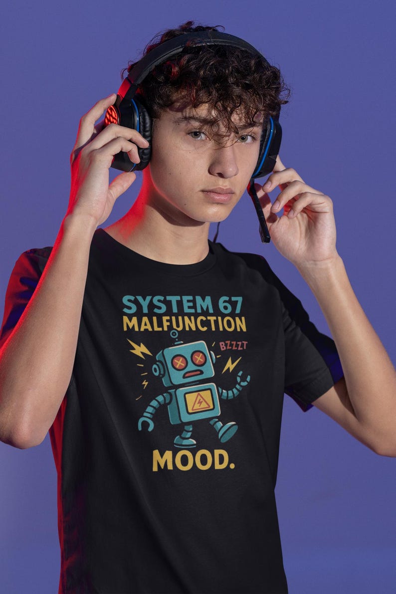 May include: Black t-shirt with a cartoon robot graphic and the text "SYSTEM 67 MALFUNCTION" and "MOOD." The robot is blue with a yellow hazard symbol. The person is wearing a black headset.