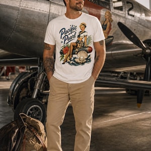 May include: A white short-sleeved t-shirt featuring a graphic print of a pin-up style woman sitting on a bomb next to a large open oyster shell with a pearl. The text Pacific Pearl is printed in blue script above the illustration.