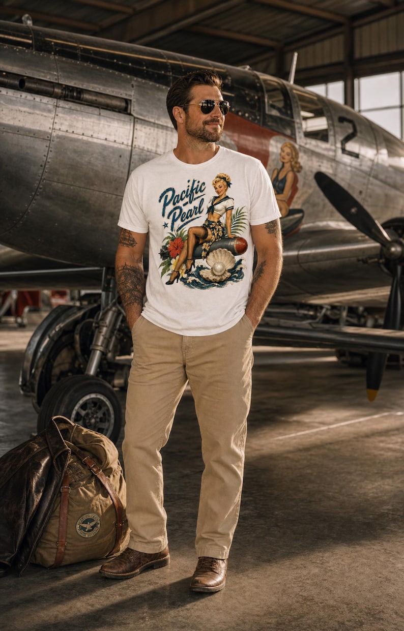 May include: A white short-sleeved t-shirt featuring a graphic print of a pin-up style woman sitting on a bomb next to a large open oyster shell with a pearl. The text Pacific Pearl is printed in blue script above the illustration.
