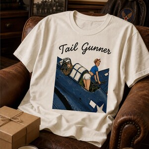 May include: A cream-colored short-sleeve t-shirt featuring a graphic print of a blue military aircraft with a pilot and a person in a blue shirt and white shorts operating a machine gun. The text Tail Gunner is printed in black script above the graphic.