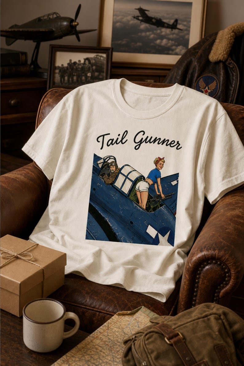 May include: A cream-colored short-sleeve t-shirt featuring a graphic print of a blue military aircraft with a pilot and a person in a blue shirt and white shorts operating a machine gun. The text Tail Gunner is printed in black script above the graphic.