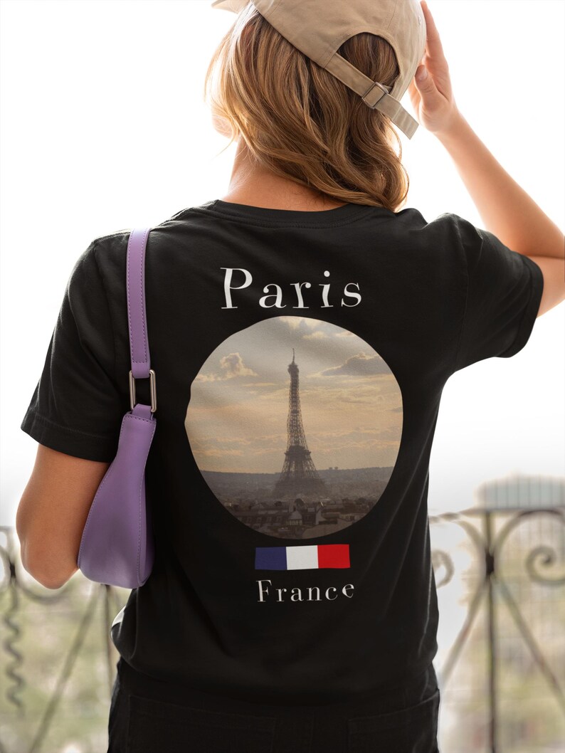 May include: Black t-shirt with a graphic of the Eiffel Tower in a circular frame, the word "Paris" above, and the French flag and the word "France" below. A purple purse hangs from the shoulder.