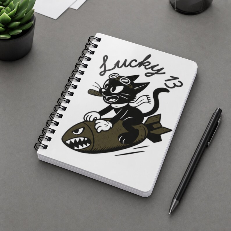 May include: A white spiral-bound notebook featuring a black and white graphic of a cartoon cat wearing goggles and a scarf while riding a bomb. The cat is smoking a cigar, and the text Lucky 13 is printed in a cursive black font above the illustration.