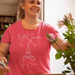 Op de afbeelding: Een koraalkleurig T-shirt met een witte afbeelding van de Eiffeltoren en de woorden "Eiffel Tower" en "City of Love". Het T-shirt heeft een ronde hals en korte mouwen. De afbeelding toont een lachende persoon. Het T-shirt is helder koraal van kleur.